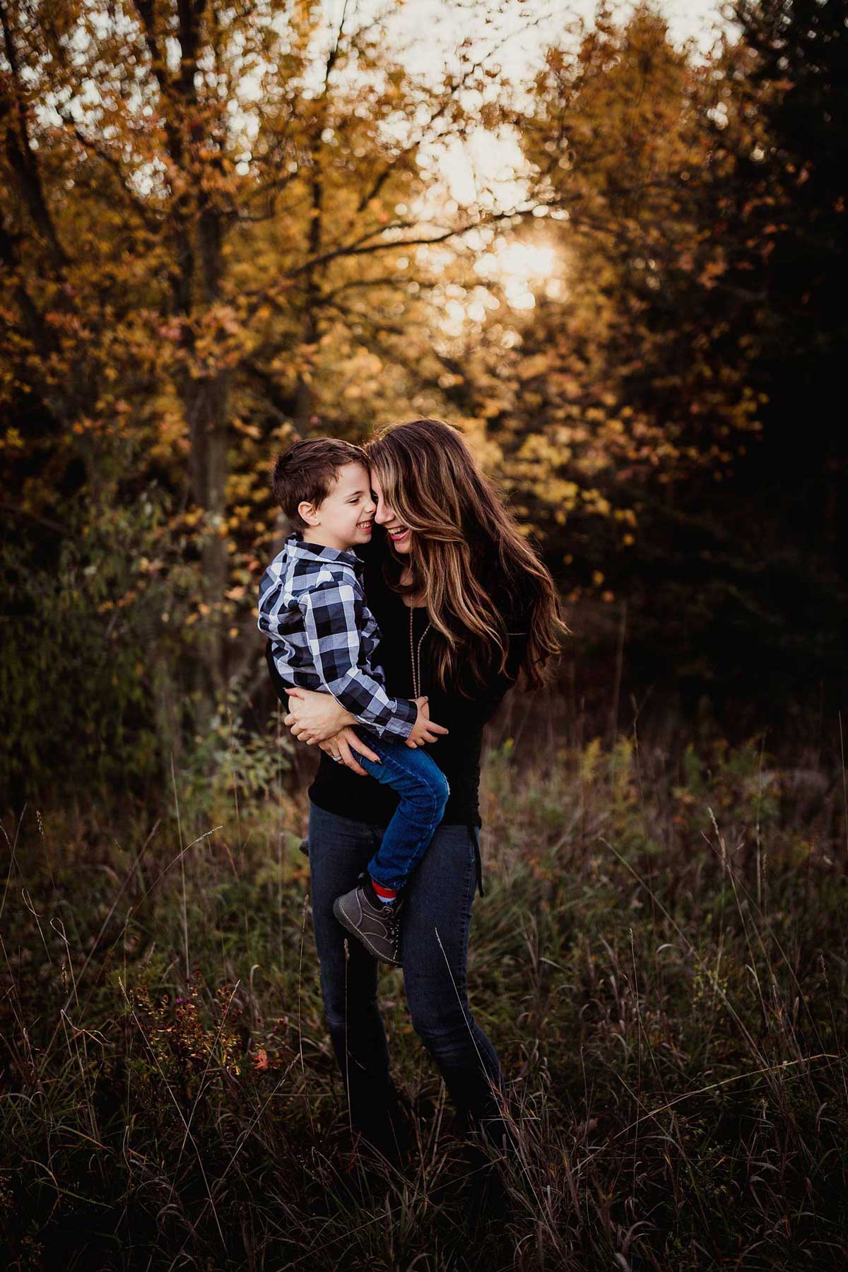Mom hugs son while holding him on her hip, with fall trees in the background.